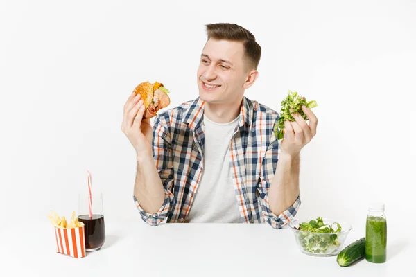 Mann sitzt am Tisch mit grünen Detox-Smoothies, Salat in Glasschüssel, Gurke, Burger, Pommes, Cola im Glas isoliert auf weißem Hintergrund. richtige Ernährung, gesunde Lebensweise, Fast Food, Auswahlkonzept. — Stockfoto
