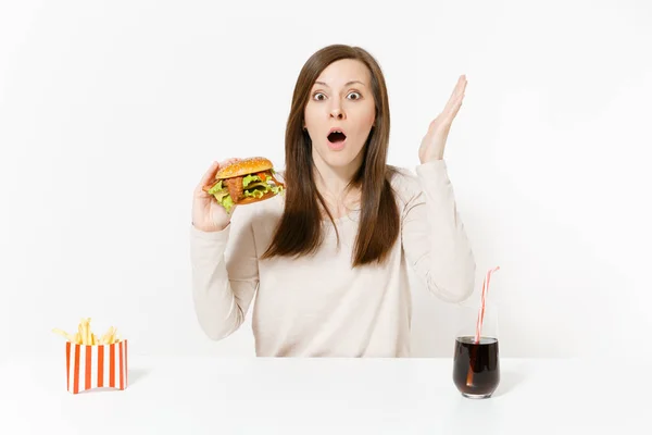 Schockierte junge Frau am Tisch mit Burger, Pommes frites, Cola in Glasflasche auf weißem Hintergrund. richtige Ernährung oder klassisches amerikanisches Fast Food. Werbefläche mit Kopierfläche. — Stockfoto