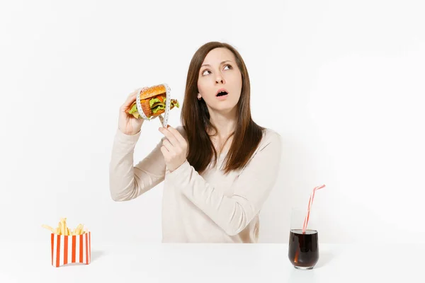 Woman at table holds burger with tailor measuring tape around, french fries, cola in glass bottle isolated on white background. Proper nutrition or American classic fast food. Area with copy space. — Stock Photo, Image