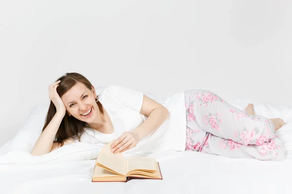 Ung glad kvinna ligger i sängen med vitt lakan, kudde, filt på vit bakgrund. Skönhet kvinna umgås i rummet, läsa bok, njuta av resten. Koppla av, gott humör koncept. Plats för text. — Stockfoto
