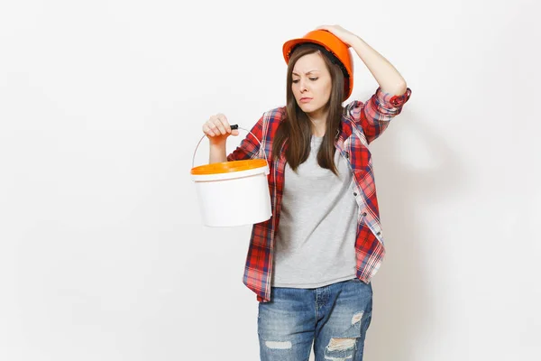 Sad dissatisfied woman in protective hardhat looking on empty paint bucket with copy space and clinging to head isolated on white background. Instrument for renovation apartment. Repair home concept. — Stock Photo, Image