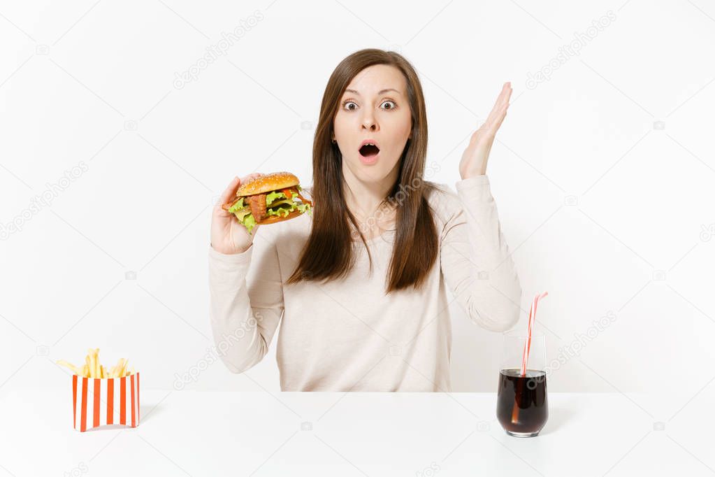 Shocked young woman sitting at table with burger, french fries, cola in glass bottle isolated on white background. Proper nutrition or American classic fast food. Advertising area with copy space.