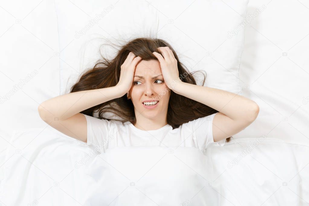 Top view of head of tired brunette young woman lying in bed with white sheet, pillow, blanket. Shocked female cover ears with hand, spending time in room. Rest, relax, good mood concept. Copy space.