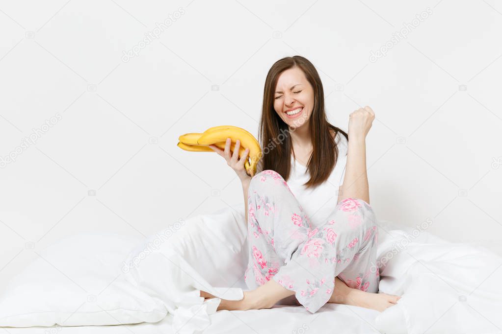 Young happy fun woman sitting in bed with white sheet, pillow, wrapping in blanket on white background. Beauty female spending time in room, holding bunch of bananas. Rest, relax, good mood concept.