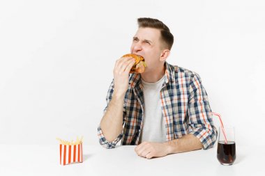 Hamburger yeme, tablo ile kızarmış patates, kola cam beyaz arka plan üzerinde izole oturup eğlenceli aç genç adam. Doğru beslenme veya Amerikan Klasik fast food. Alan kopya alanı ile reklam