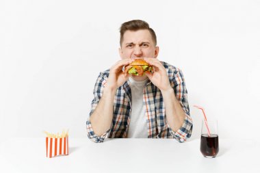 Hamburger yeme, tablo ile kızarmış patates, kola cam beyaz arka plan üzerinde izole oturup eğlenceli aç genç adam. Doğru beslenme veya Amerikan Klasik fast food. Alan kopya alanı ile reklam