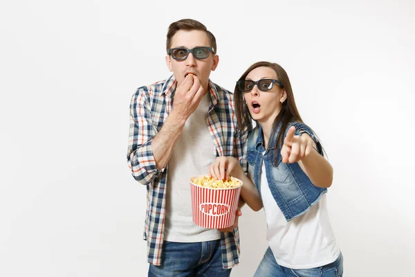 Young shocked couple, woman and man in 3d glasses watching movie film on date, holding bucket of popcorn, stealing popcorn, pointing index finger isolated on white background. Emotions in cinema