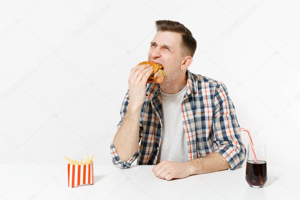 Fun hungry young man eating burger, sitting at table with french fries, cola in glass isolated on white background. Proper nutrition or American classic fast food. Advertising area with copy space