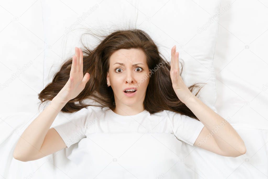 Top view of head of tired brunette young woman lying in bed with white sheet, pillow, blanket. Shocked female cover ears with hand, spending time in room. Rest, relax, good mood concept. Copy space