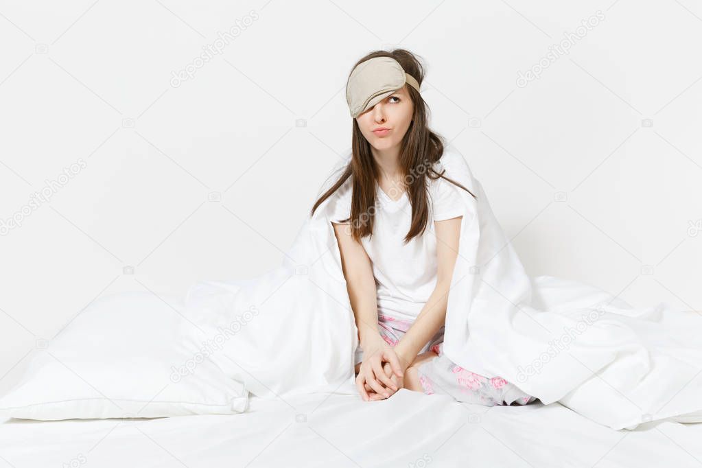 Fun tired young woman sitting in bed with sleep mask, sheet, pillow, wrapping in blanket isolated on white background. Beauty female spending time in room. Rest, relax, good mood concept. Copy space
