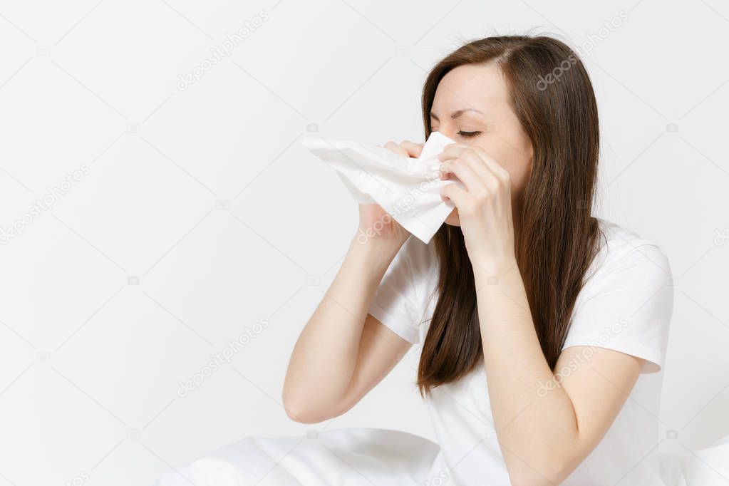 Young sickness tired brunette woman in pajamas sitting on bed isolated on white background. Female feeling bad, blowing her nose into white napkin. Cold in the head. Copy space for advertisement