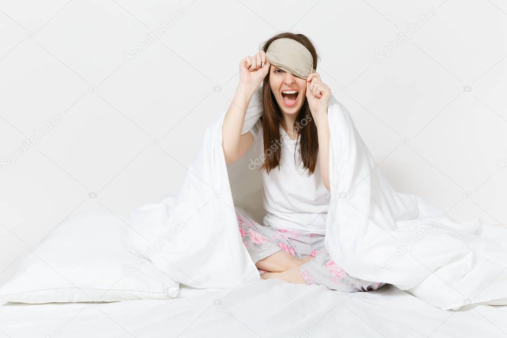 Fun tired young woman sitting in bed with sleep mask, sheet, pillow, wrapping in blanket isolated on white background. Beauty female spending time in room. Rest, relax, good mood concept. Copy space