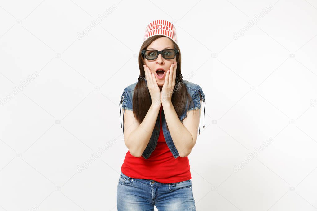 Portrait of young shocked attractive woman in 3d glasses and casual clothes with bucket for popcorn on head watching movie film and clinging to face isolated on white background. Emotions in cinema