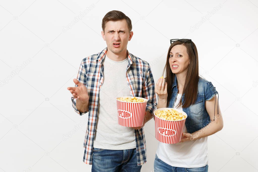 Young irritated dissatisfied couple, woman and man in 3d glasses and casual clothes watching movie film on date, holding buckets of popcorn isolated on white background. Emotions in cinema concept