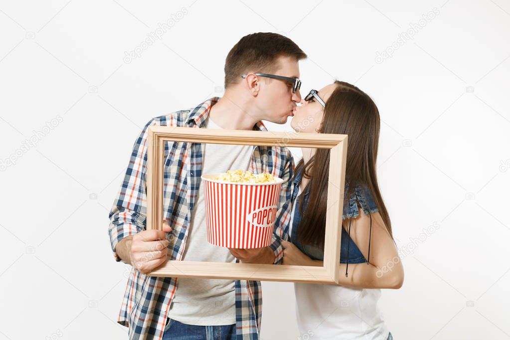 Young kissing couple, woman and man in 3d glasses and casual clothes watching movie film on date, holding bucket of popcorn and picture frame isolated on white background. Emotions in cinema concept