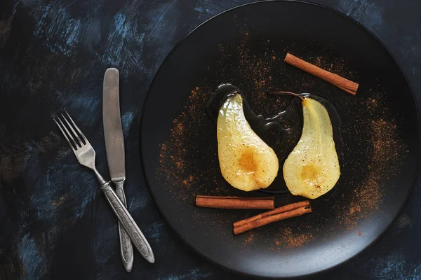 Pear poached with cinnamon sticks served a plate, fork, knife, on a blue background.