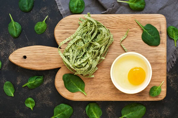 Dry green noodles with spinach and raw egg in a bowl on a dark background, top view