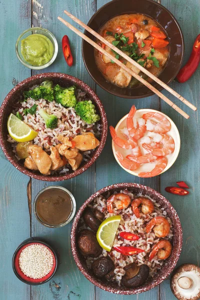 A variety of dishes with Asian food, brown rice, fried shrimp, fried mushrooms, braised chicken, sauces. Green-blue rustic background. Close-up, top view.