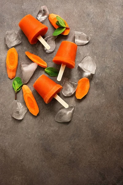 Homemade carrot popsicles with crushed ice and mint leaves on a dark rustic background. Vegan and vegetarian dessert. Frozen juice. Top view, flat lay, copy space