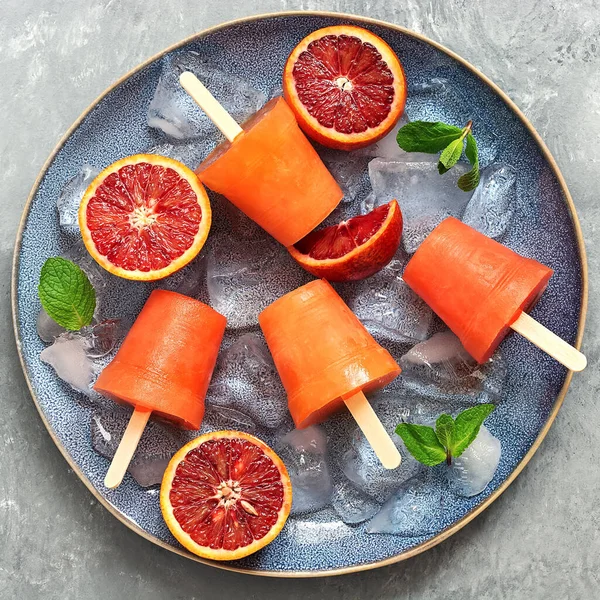 Frozen juice of red orange on a stick with ice and slices of fruit. Ice cream popsicles. Summer refreshing dessert. Top view, flat lay, square.