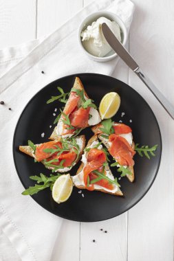 Somon, peynir, roka ve limonlu sandviç. Beyaz bir masada, siyah bir tabakta.