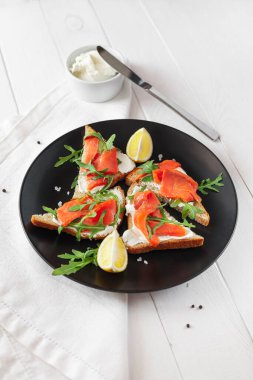 Somon, peynir, roka ve limonlu sandviç. Beyaz bir masada, siyah bir tabakta.