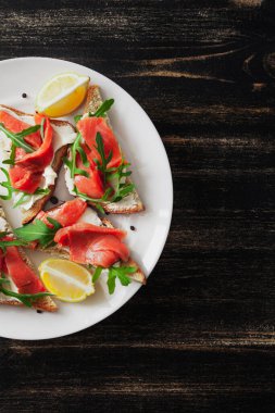 Somon, peynir, roka ve limonlu sandviç siyah bir masada beyaz tabakta.