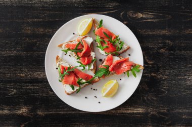 Somon, peynir, roka ve limonlu sandviç siyah bir masada beyaz tabakta.