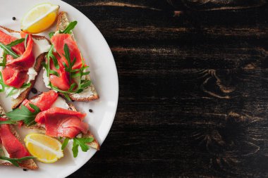 Somon, peynir, roka ve limonlu sandviç siyah bir masada beyaz tabakta.