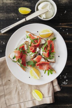 Somon, peynir, roka ve limonlu sandviç beyaz bir tabakta keten peçete ve siyah bir masa üzerinde malzemelerle.