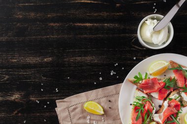 Somon, peynir, roka ve limonlu sandviç beyaz bir tabakta keten peçete ve siyah bir masa üzerinde malzemelerle.