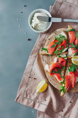 Somon, peynir, roka ve limonlu sandviç ahşap bir tahtanın üzerinde keten peçete ve gri bir masada malzemelerle.