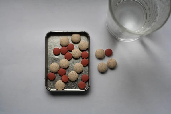 Handful of beige and red pills in steel tablet box on the table next to a glass of water