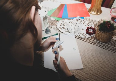 Kız yılbaşını ya da yeni yılı kutlamak için kağıttan kar taneleri kesiyor. Beyaz ve renkli kağıt, makas, keçeli kalem, beyaz dantel masa örtüsü üzerindeki sezon dekorasyonu