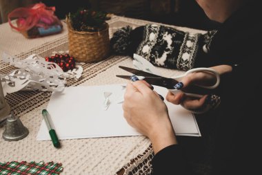 Kız yılbaşını ya da yeni yılı kutlamak için kağıttan kar taneleri kesiyor. Beyaz ve renkli kağıt, makas, keçeli kalem, beyaz dantel masa örtüsü üzerindeki sezon dekorasyonu