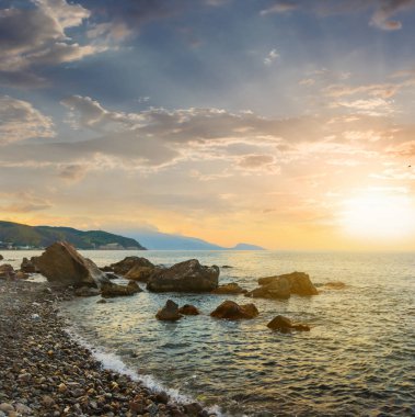 Gün batımında kıyıya yakın taşlarla sakin bir deniz koyunda, açık havada tatil sahnesi.