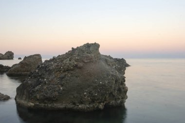Alacakaranlıktaki deniz kıyısında büyük kayalar, sakin bir deniz koy arka planı.