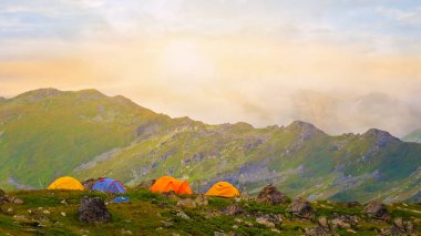 Gün batımında yeşil bir dağ sırtı yakınında turistik kamp, doğa yürüyüşü sahnesi.