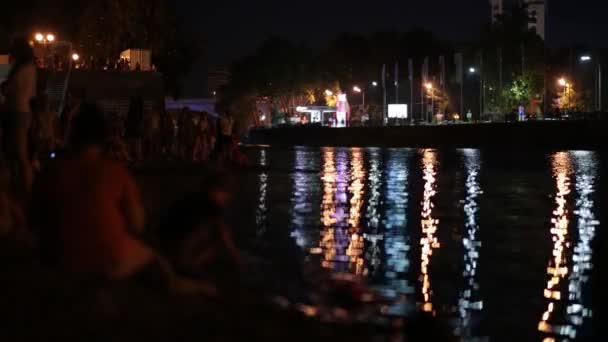 Gente en la playa por la noche. Luces reflejos en el agua . — Vídeos de Stock