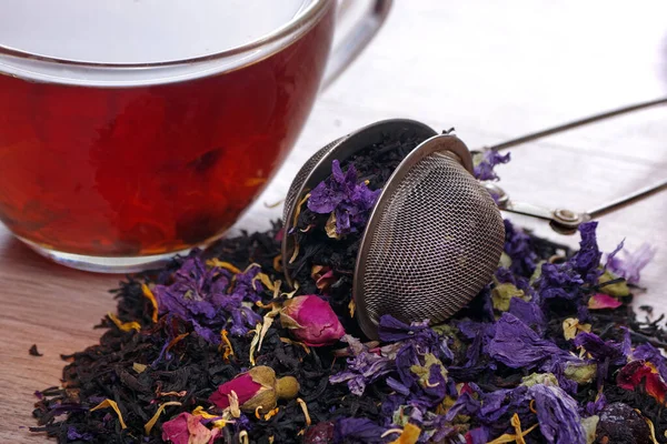 heap of blended tea with dry roses and flower petals, tea strainer and cup of tea on a wooden table