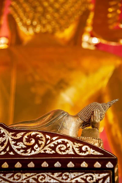 STILL LIFE OF RECLINING BUDDHA STATUE. Bronze Buddha statue in reclining posture with right hand supporting the head, lying on a rosewood bed with pearl inlay. In front, there is another Buddha statue out of focus.