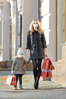 Kırmızı alışveriş çantalı, tıbbi maskeli bir kadın ve küçük bir çocuk sokakta yürüyor. Covid-19 koronavirüs salgınını hesapla