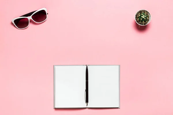 Diario en blanco abierto, bolígrafo negro, gafas blancas sobre fondo de texto rosa. plano, vista superior. Espacio de trabajo femenino — Foto de Stock