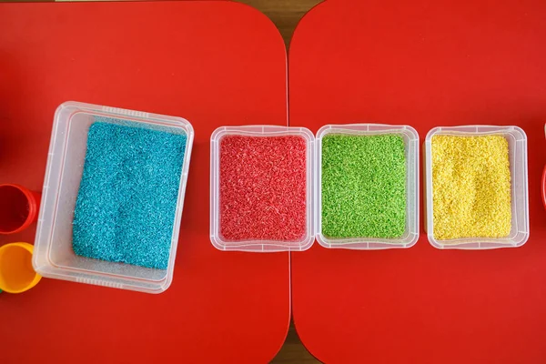 Coș de gunoi senzorial pentru copiii mici cu orez colorat pe masa roșie. Joc educațional universal — Fotografie, imagine de stoc