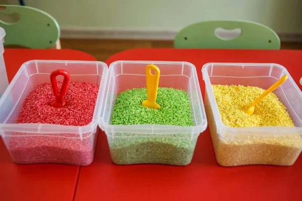 Сенсорная корзина для малышей с разноцветным рисом на красном столе . Лицензионные Стоковые Изображения