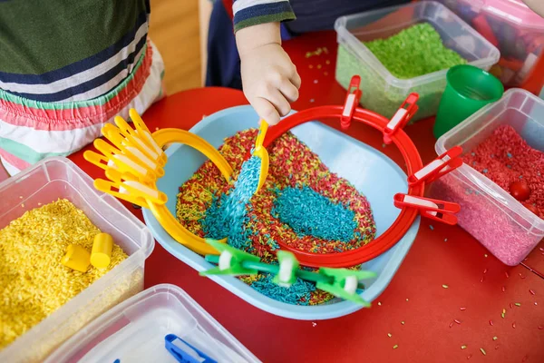 Toddlers bermain dengan tempat sampah sensorik dengan beras warna-warni di atas meja merah . Stok Lukisan  