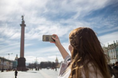 Young traveler woman wearing casual classic coat, holding cellphone and taking photo Alexandrian Column while walking in Saint-Petersburg, Palace Square in spring or autumn time clipart