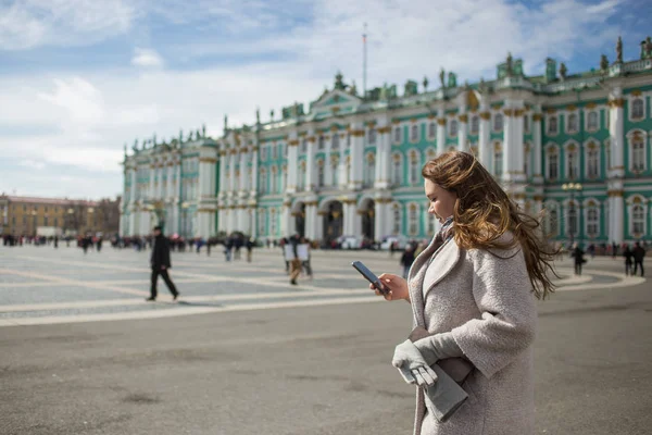 Молодая путешественница в классическом пальто, с мобильным телефоном, с помощью приложения, GPS, ищет направление во время прогулки по Санкт-Петербургу, Дворцовой площади, Зимнему дворцу весной или осенью — стоковое фото
