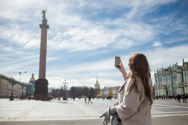 Молодая путешественница, одетая в классическое пальто, держит мобильный телефон и фотографирует Александрианскую колонну во время прогулки по Санкт-Петербургу, Дворцовой площади весной или осенью — стоковое фото