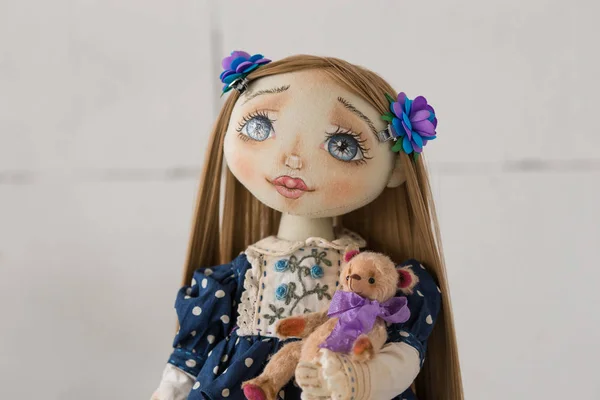 Portrait of textile handmade vintage doll with blue eyes, long brown hair in old blue textile dress with gentle print, in white shirt on white background. She holding little textile teddy bear